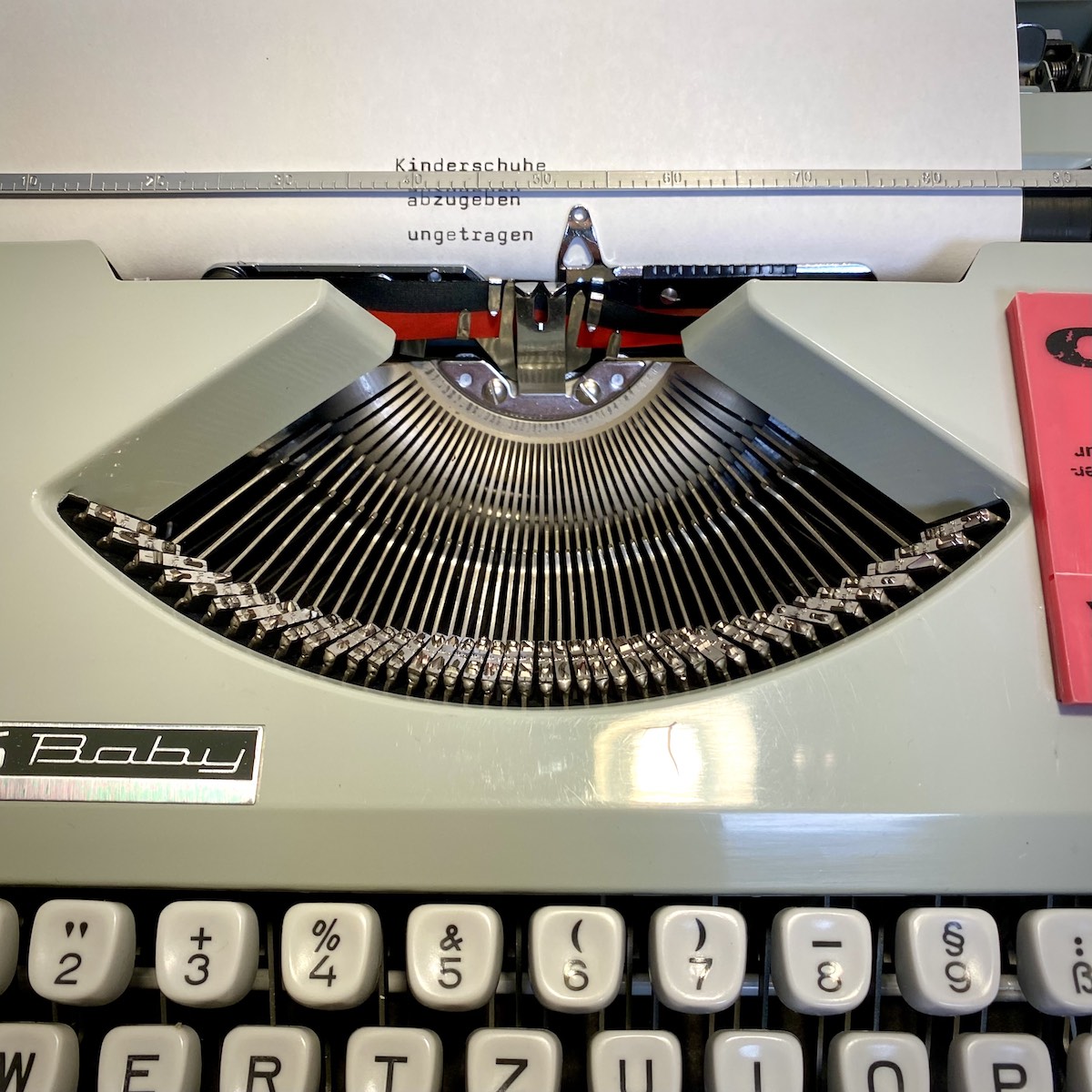 Ein Bildausschnitt einer mechanischen Schreibmaschine Typ Hermes Baby, darin ein eingespanntes Blatt, auf das die Worte Kinderschuhe abzugeben ungetragen getippt sind